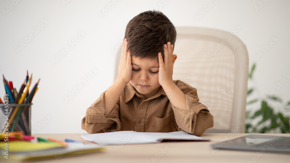 Unhappy sad tired caucasian little child sits at table presses hands to head, suffers from tension and difficult Unhappy sad tired caucasian little child sits at table presses hands to head, suffers from tension and difficult