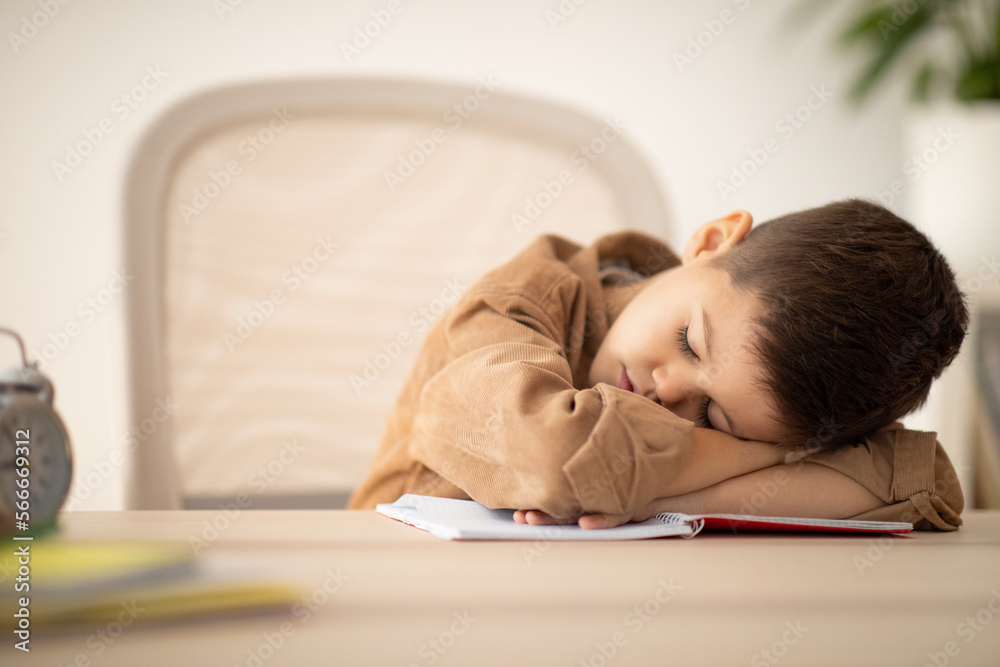 Tired small european boy lies at table sleeping, resting from lesson in school room interior Tired small european boy lies at table sleeping, resting from lesson in school room interior