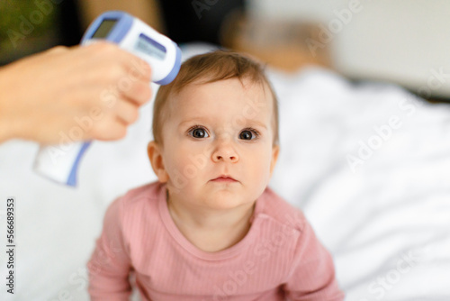 Closeup portrait of mother measuring temperature of her adorable baby girl with infrared thermometer, sitting on bed