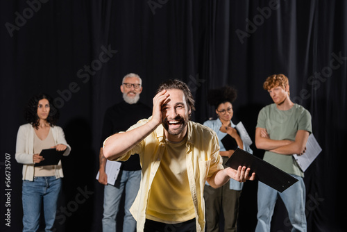 laughing man touching face and looking at camera near blurred multiethnic actors and producer in theater school.