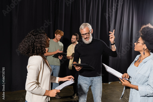 bearded art director pointing with finger while reading screenplay near multiethnic actresses during acting skills lesson.