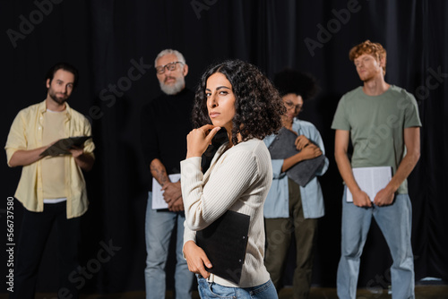 multiracial woman with strict face expression looking at camera while rehearsing near art director and actors on blurred background.