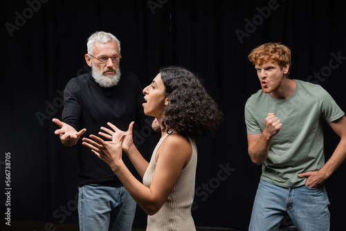multicultural actress rehearsing near gesturing acting skills teacher and emotional redhead man in theater.