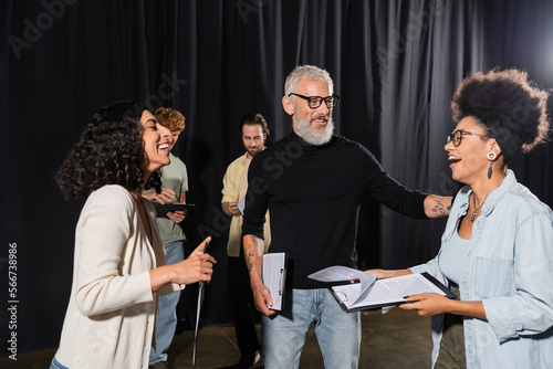 cheerful african american woman holding screenplay and laughing near bearded producer and multiethnic actors in theater.