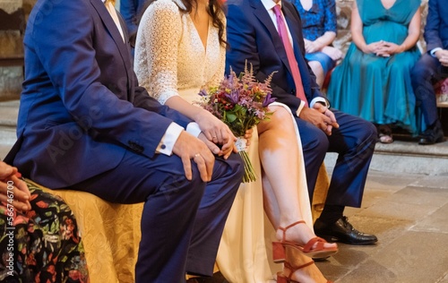 Newly married couple sitting in the church pew while holding hands where rings are visible. Also seen in the image is an unrecognizable person who is the best man at the wedding.