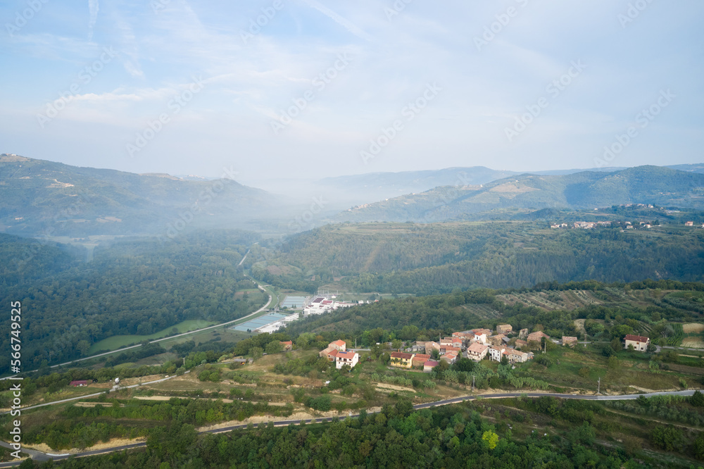 A mountainous, forested landscape with villages, vegetable gardens and vineyards in the morning mist. A winding road runs down the mountain plateau into the distance. Shot from a drone.
