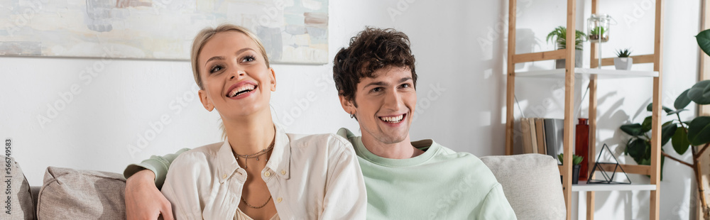 happy and curly young man hugging blonde girlfriend at home, banner.