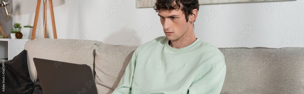 curly young man using laptop while sitting on couch in living room, banner. curly young man using laptop while sitting on couch in living room, banner.