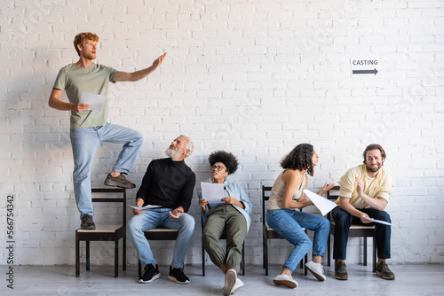 Φωτογραφία redhead man standing on chair and rehearsing near multiethnic actors sitting and waiting for casting