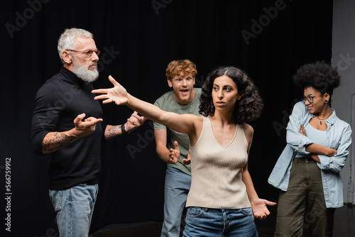 multiracial woman standing with outstretched hand near bearded art director and emotional interracial students in acting school.