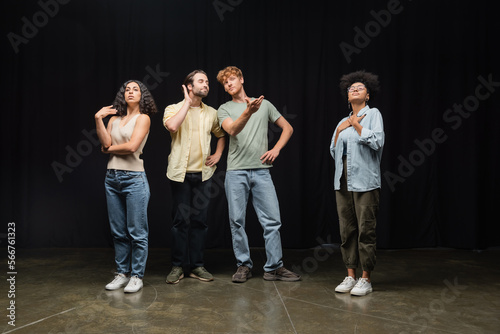 redhead man standing with hands on hips and pointing with hands near multicultural actors in acting studio.