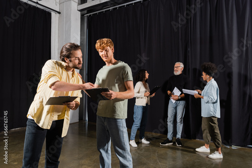young actors holding clipboards with screenplays near interracial theater troupe on background.