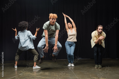 excited redhead man jumping near multiethnic actors posing during rehearsal in theater. photo