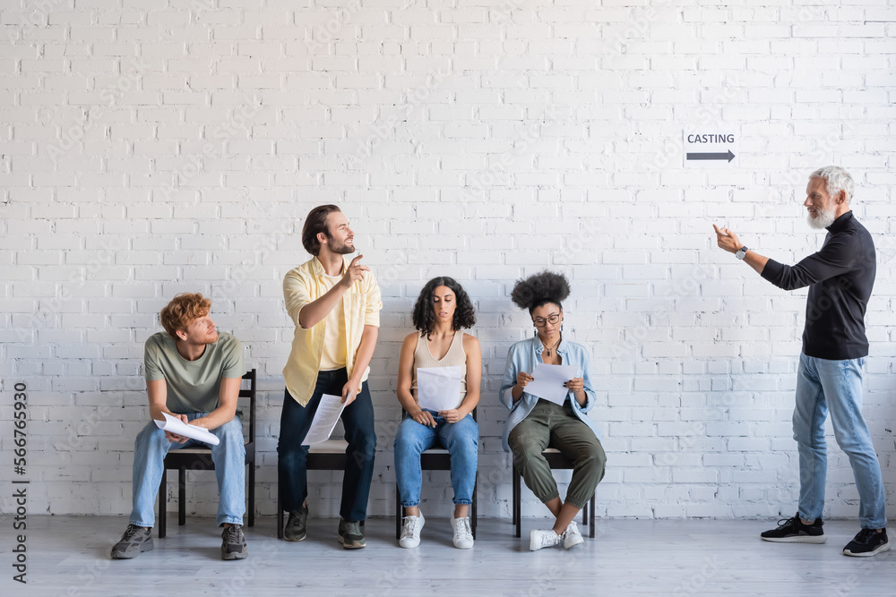 Fototapeta premium bearded art director calling brunette man near multiethnic actors sitting on chairs and waiting for casting.