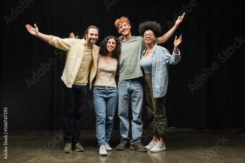 full length of cheerful interracial actors embracing while waving hands and smiling at camera in theater.