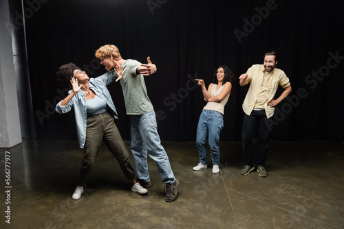 cheerful interracial students pointing at redhead man and african american woman rehearsing on theater stage.