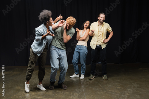 cheerful interracial actors pointing at african american woman rehearsing with redhead man on stage in theater.