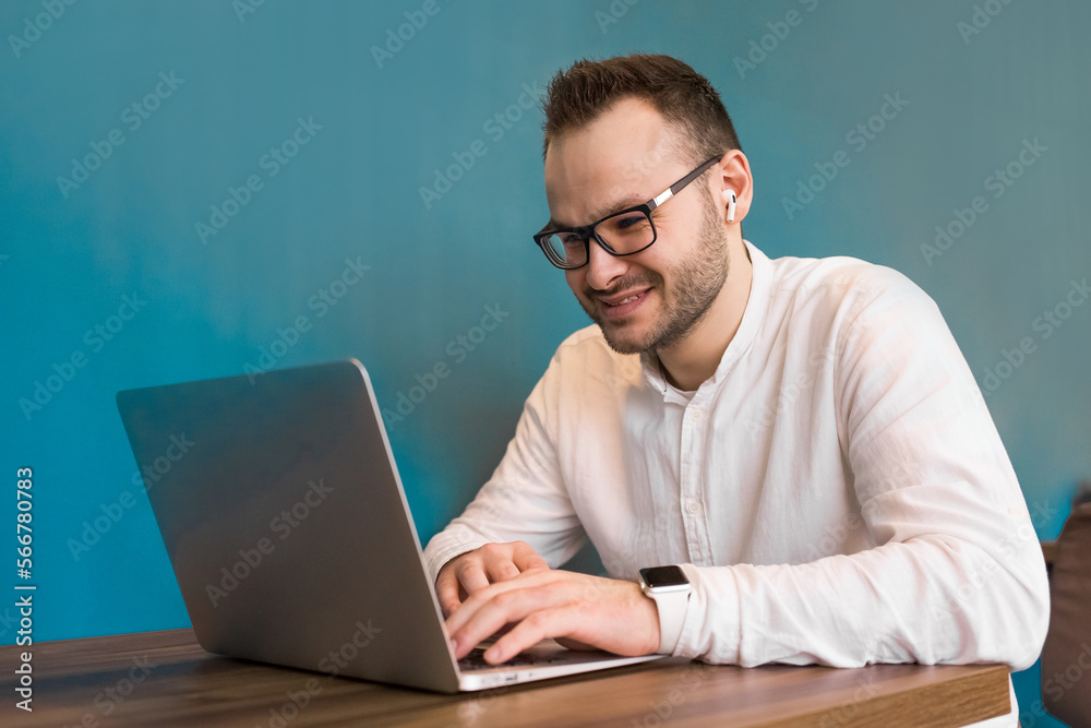 A young attractive businessman guy with a beard and glasses works in a laptop sitting at a table in a cafe and smiling A young attractive businessman guy with a beard and glasses works in a laptop sitting at a table in a cafe and smiling