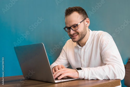 A young attractive businessman guy with a beard and glasses works in a laptop sitting at a table in a cafe and smiling © Andrey