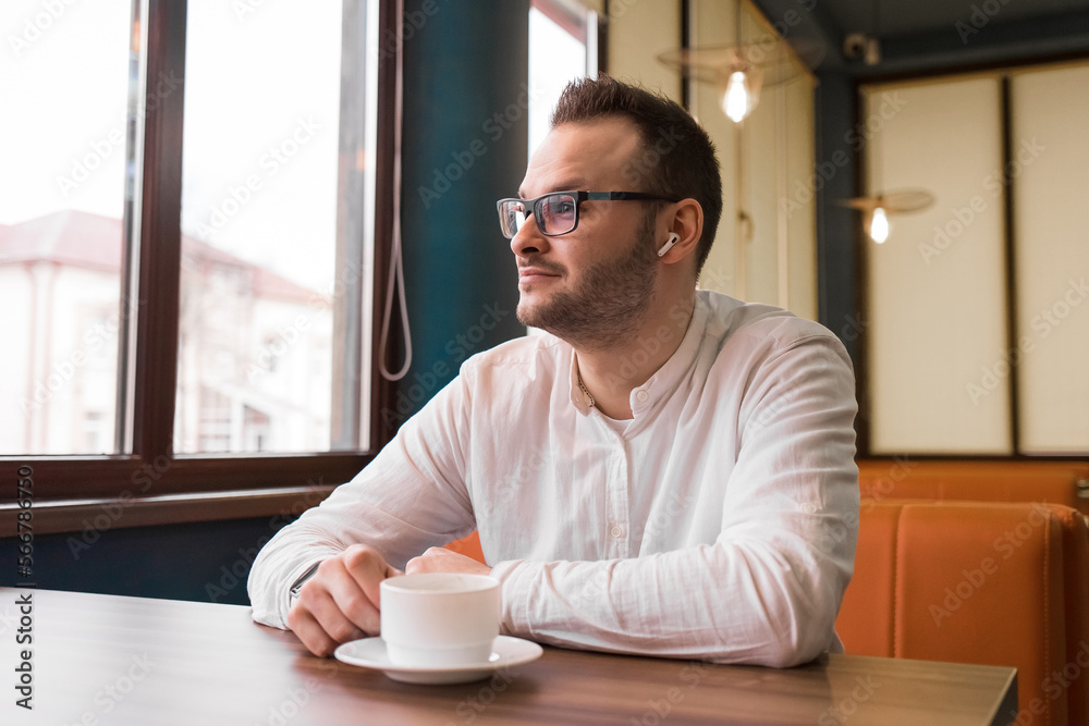 Young attractive businessman in a white shirt, glasses and headphones drinks coffee and looks thoughtfully out the window Young attractive businessman in a white shirt, glasses and headphones drinks coffee and looks thoughtfully out the window