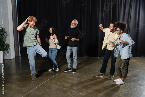 redhead man posing near excited multicultural actors and grey haired producer on stage in acting skills school.