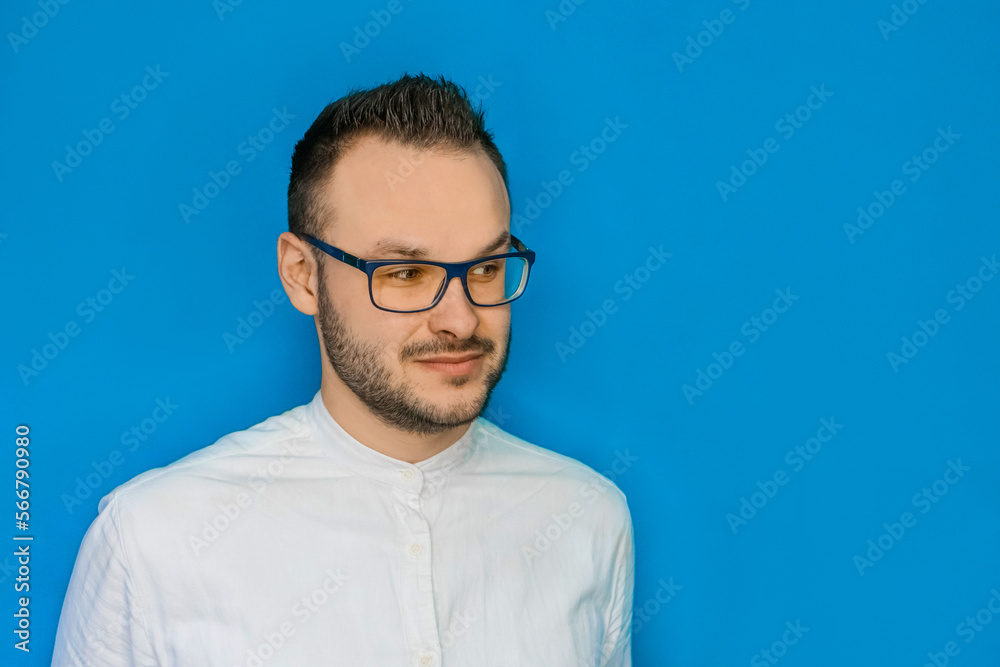 Portrait of a stylish young attractive guy with glasses and a white shirt looking to the side against a blue background