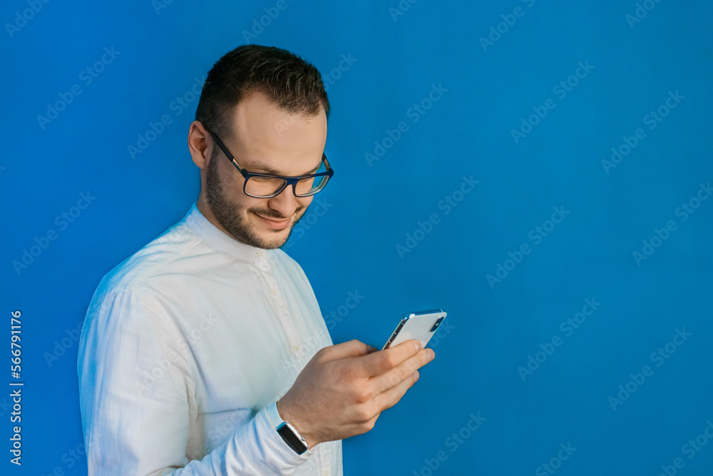 Portrait of a cheerful stylish young guy businessman Caucasian appearance in glasses and white shirt, holds a smartphone in his hand and spends time in it on a blue background