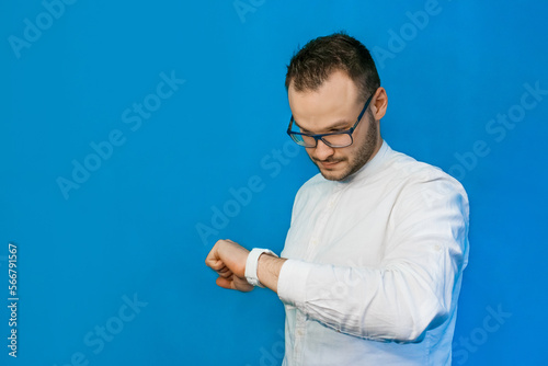 Stylish, business-like young portrait guy Caucasian-looking businessman in glasses and white shirt, looks time on smart clock on blue background