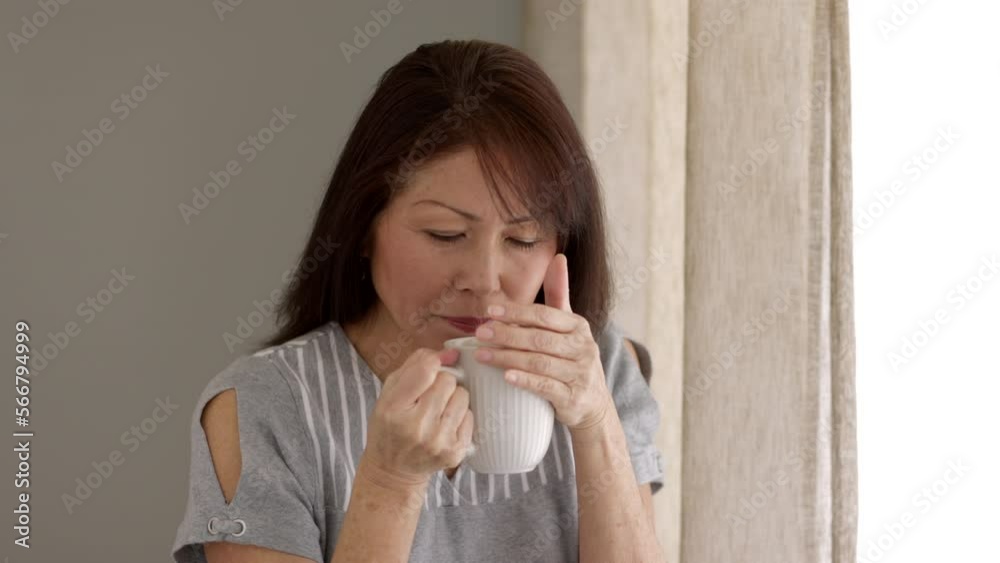 Attractive middle age woman looking at camera relaxing drinking coffee sitting near window in natural leisure home living room Attractive middle age woman looking at camera relaxing drinking coffee sitting near window in natural leisure home living room