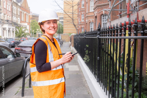 Female chartered civil engineer taking notes on her smart phone with am electronic pen, construction site, hard hat and orange high visibility vest, inspection, smiley, happy, looking at camera