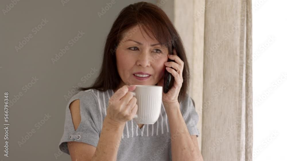 Attractive middle age woman relaxing talking on mobile phone drinking coffee sitting near window in natural leisure home living room Attractive middle age woman relaxing talking on mobile phone drinking coffee sitting near window in natural leisure home living room