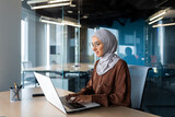 Successful smiling Arab woman in hijab working inside modern office, Muslim woman using laptop at work, business woman satisfied with achievement results typing on computer keyboard.