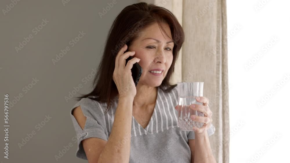 Attractive mature woman relaxing talking on mobile phone drinking water sitting in natural leisure home living room near window Attractive mature woman relaxing talking on mobile phone drinking water sitting in natural leisure home living room near window