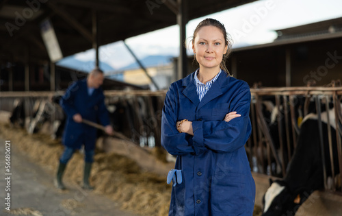 Positive woman farmer standing in cowshed with arms crossed and smiling.