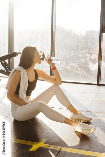 Relaxing after training. beautiful young woman looking away while sitting at gym and drink water. Young female at gym taking a break from workout. © dsheremeta