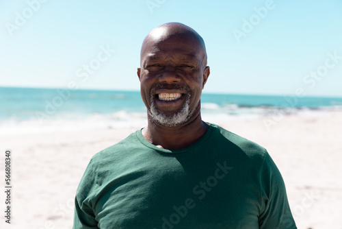 Portrait of smiling african american bald senior man standing at beach against sea and clear sky