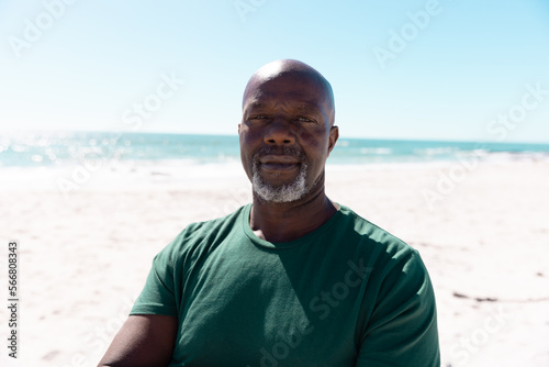Portrait of confident bald african american senior man standing at beach against sea and clear sky