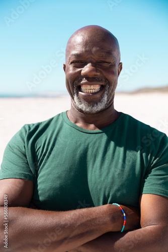 Carefree bald african american senior man with arms crossed smiling at beach against clear sky