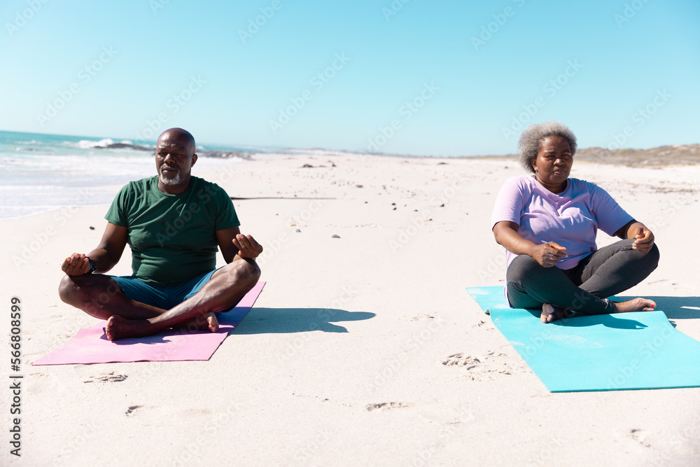 African american senior couple meditating while sitting on exercise mat at beach under clear sky African american senior couple meditating while sitting on exercise mat at beach under clear sky