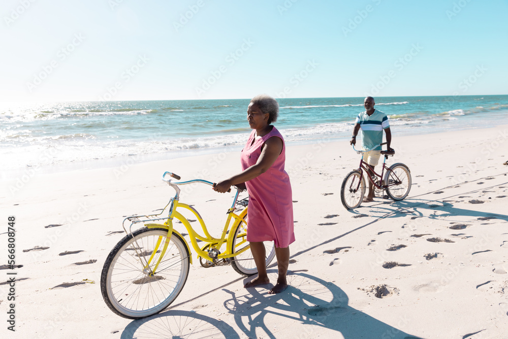 African american senior couple with bikes walking on sandy beach against sea and clear sky in summer