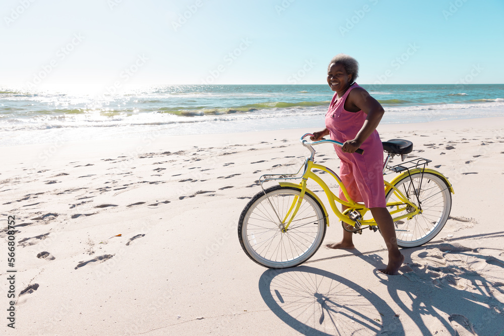 Carefree african american senior woman with bicycle standing on sandy beach against clear sky