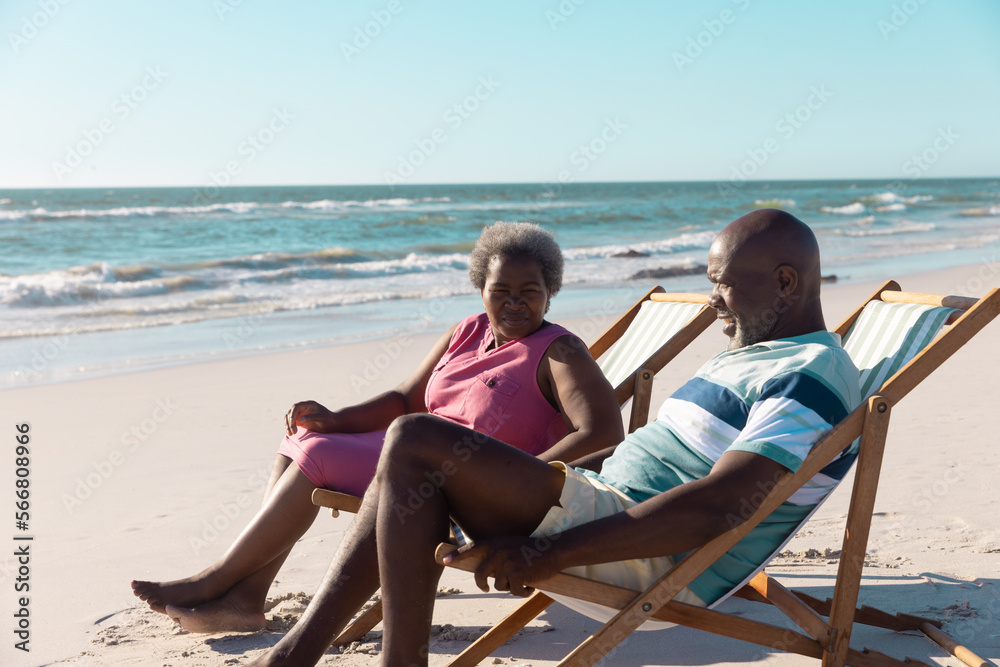 African american senior couple talking while relaxing on deckchairs against seascape and clear sky
