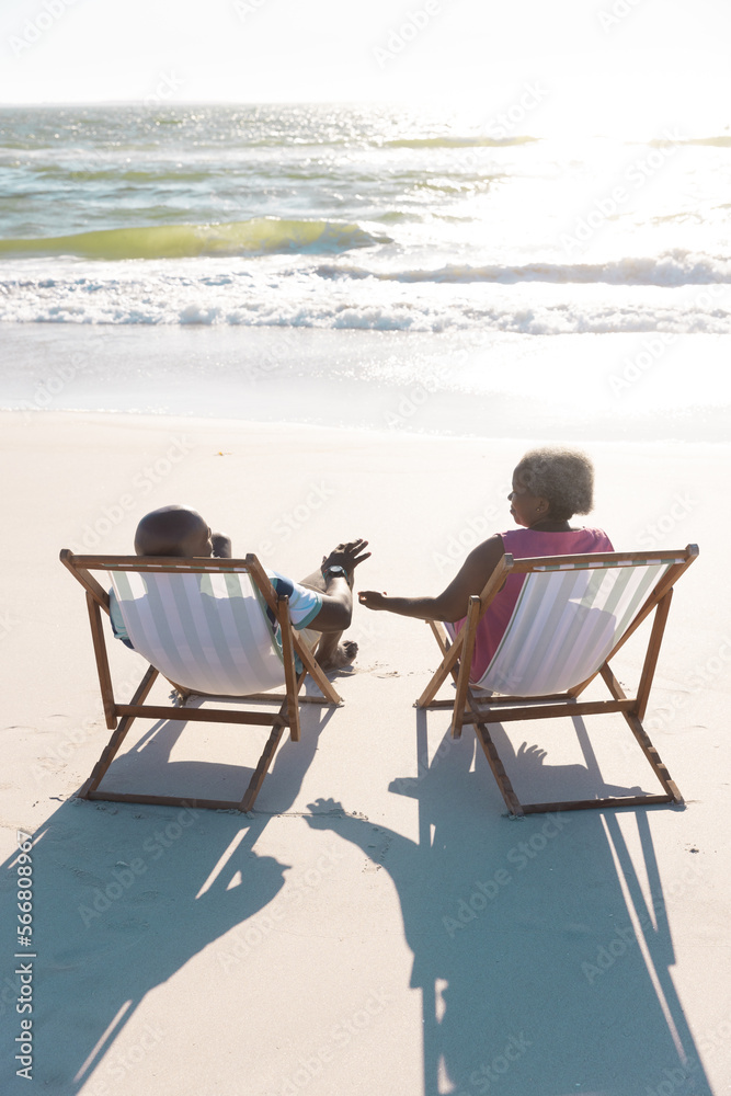 Rear view of african american senior couple sitting on deckchairs at beach in front of sea under sky