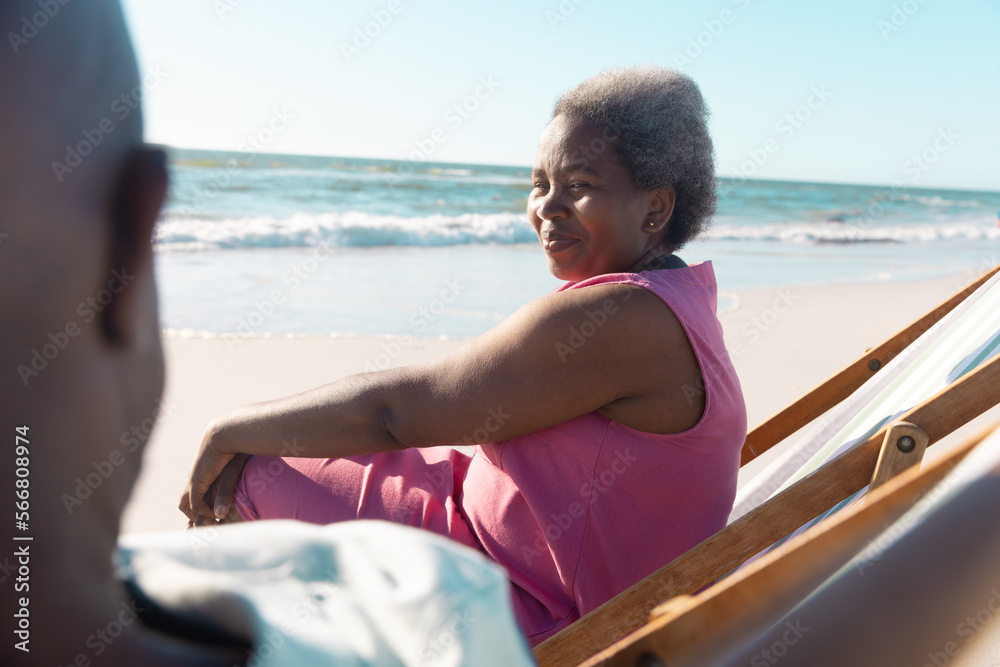Smiling african american senior woman talking with man while relaxing on deckchairs at beach