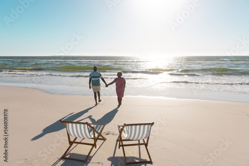 Rear view of african american senior couple holding hands and walking towards sea under sky