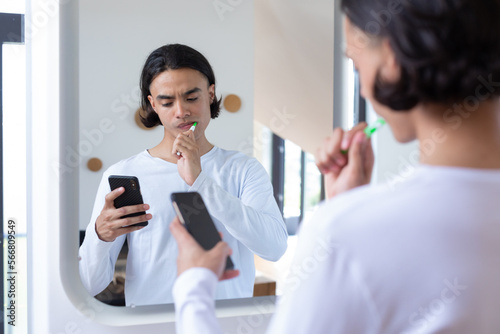 Happy biracial man brushing teeth and using smartphone in bathroom