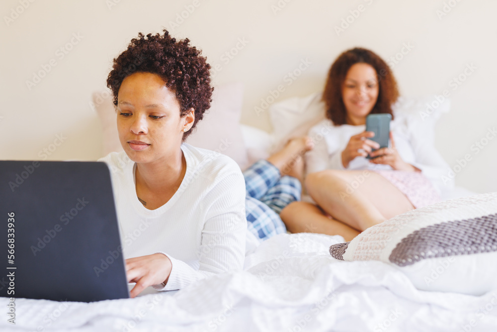 Happy diverse lesbian couple lying in bed, using laptop and smartphone Happy diverse lesbian couple lying in bed, using laptop and smartphone