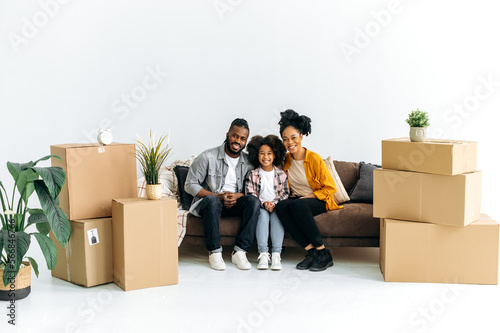 Photo of a happy African American family, joyful dad mom and their preschool daughter, sit on the sofa in the living room between boxes with things, moved to new housing, look at the camera, smile © Kateryna