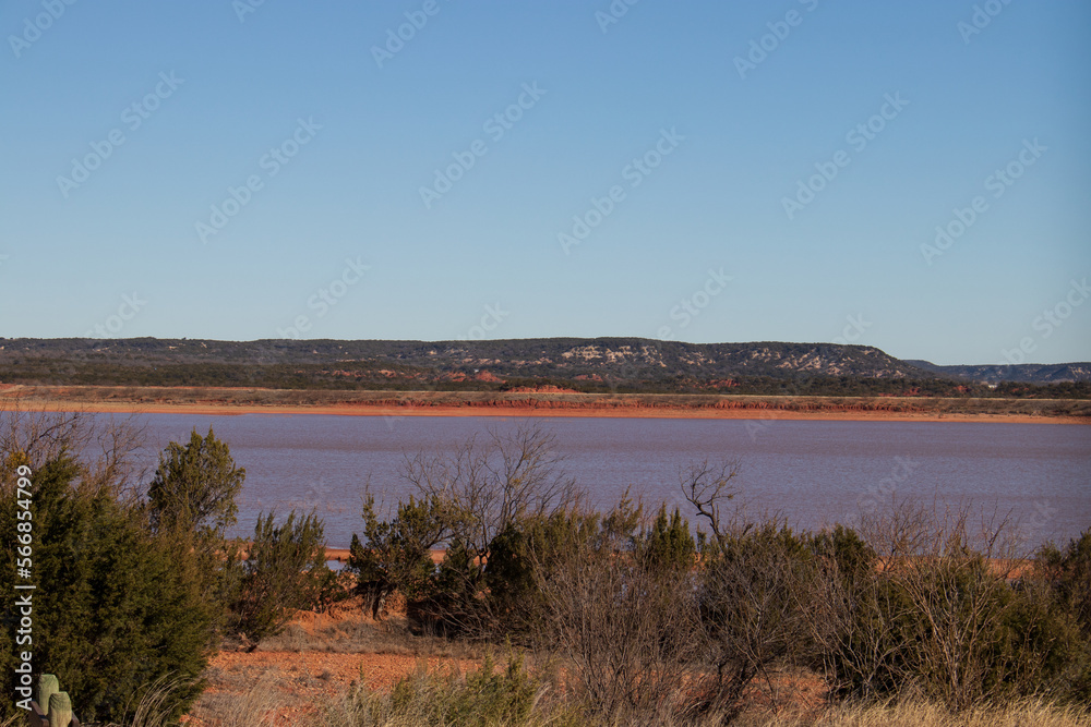 A view of Lake Abilene with the red clay shoreline and the rolling hills of Texas in the background at Abilene State Park, Texas