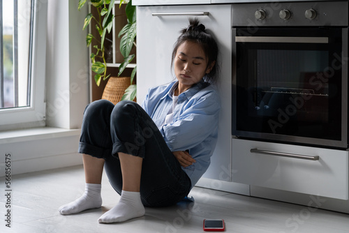 Offended unhappy Asian woman sadly looking at phone sits on floor at kitchen. Sad thoughtful worried Korean girl experiencing depression after breaking up with beloved man or hurtful social media post photo